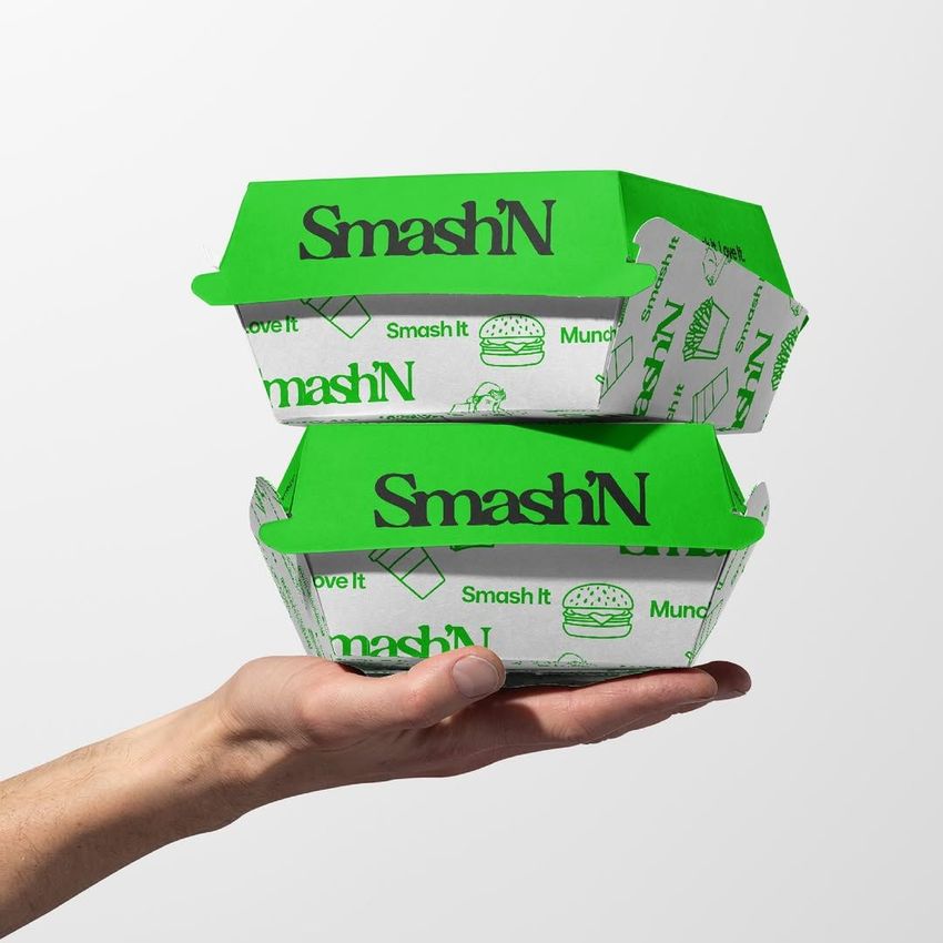 Packaging mockup featuring two green and white burger boxes with bold text, stacked on a hand against a plain background.