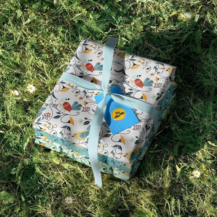 Fabric stack mockup featuring folded textiles with a spring bird pattern, tied with a light blue ribbon, placed on grass with daisies.