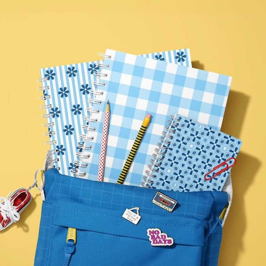 School backpack mockup with spiral notebooks in blue patterns, pencils, and a shoe keychain on a yellow background.