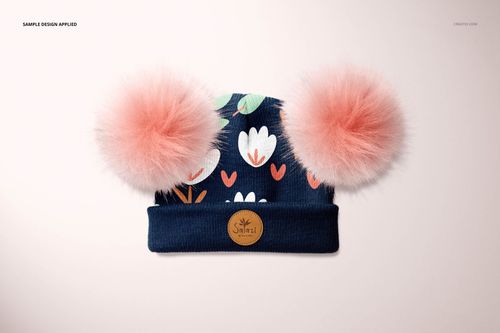 PSD mockup showing a children's beanie with fluffy pink pompons, dark blue color, and white flower design, top view.