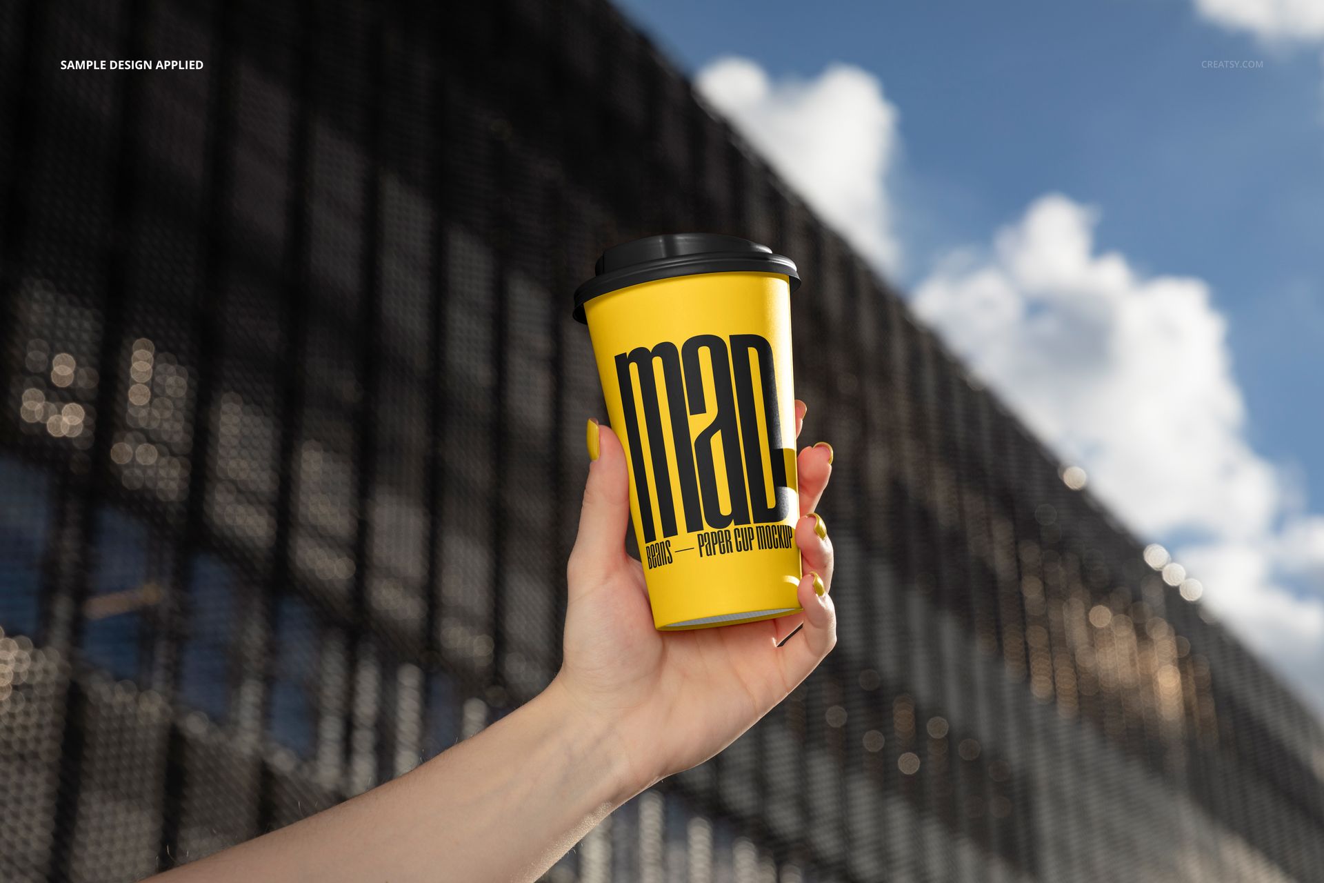 PSD mockup of a paper cup in yellow with black typography, held up outdoors with a building and blue sky in the background.
