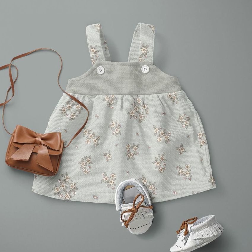 Baby dress mockup displayed flat on a gray surface, featuring a subtle flower pattern, with white shoes and a brown purse.
