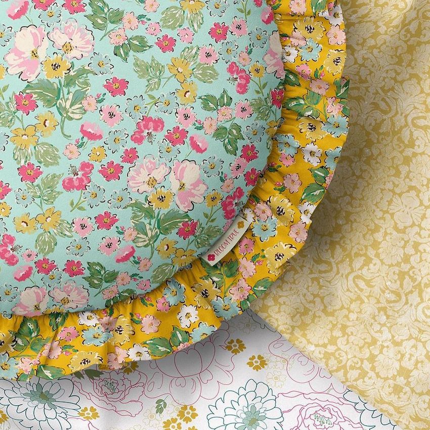 Ruffled pillow mockup featuring a round cushion with a blue floral pattern and yellow ruffled edge, placed on patterned fabrics.