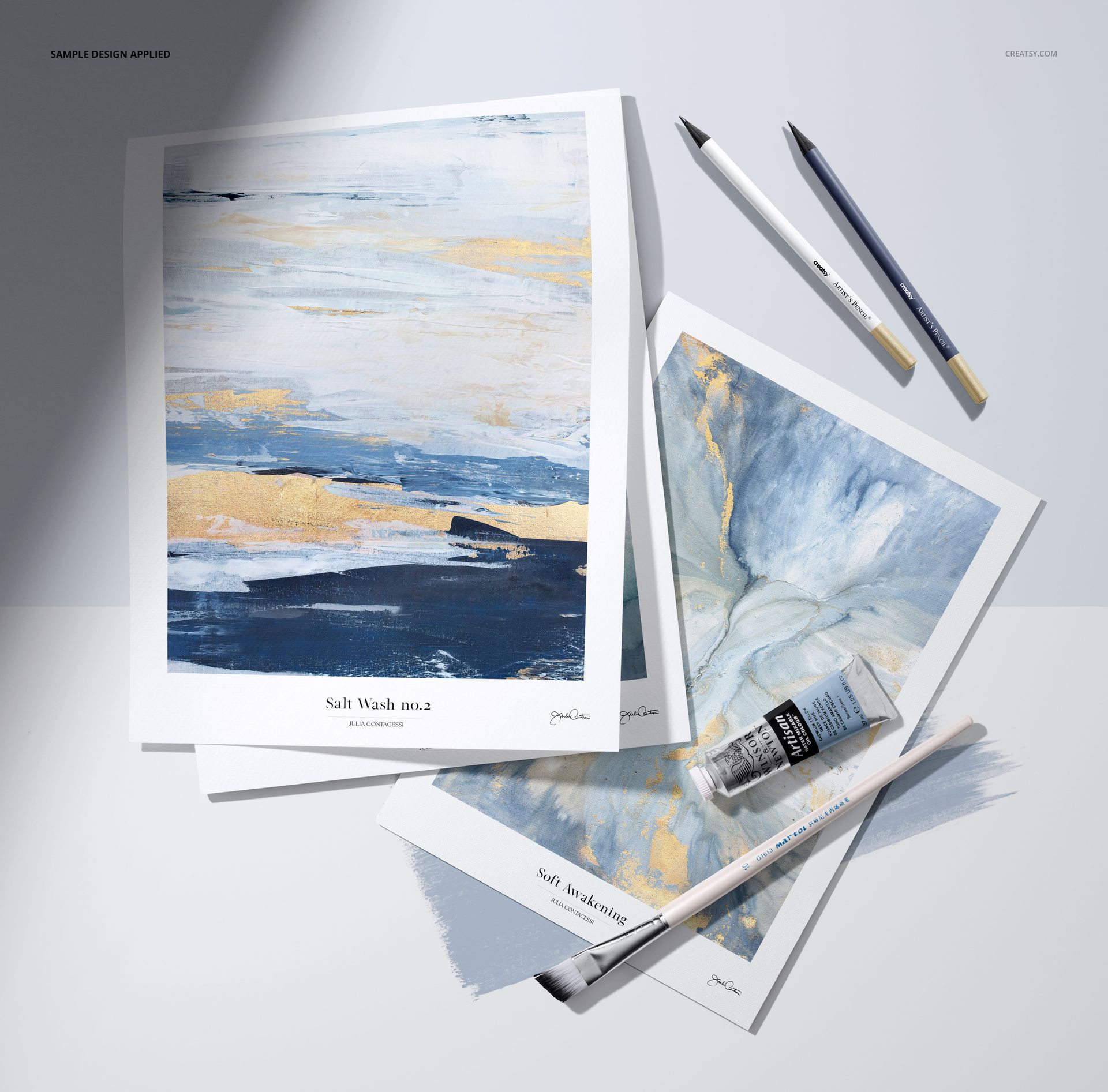 PSD mockup showing two paper pads featuring abstract art in blue and gold tones, surrounded by art supplies and pencils.