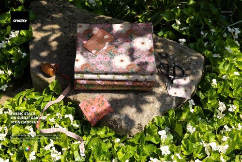 PSD mockup showing several folded fabrics with pink and white flower patterns on a garden rock, with sewing tools and greenery.