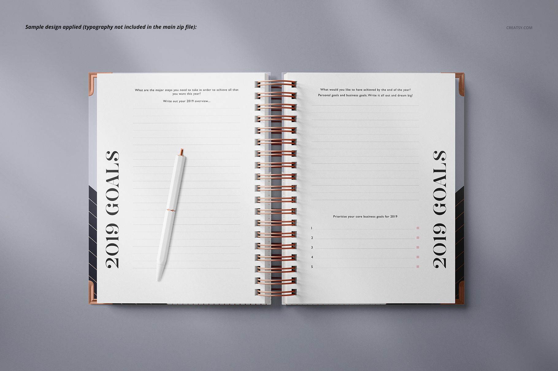 Wire-bound planner mockup with open pages, copper spiral binding, and a white pen on a light gray surface.