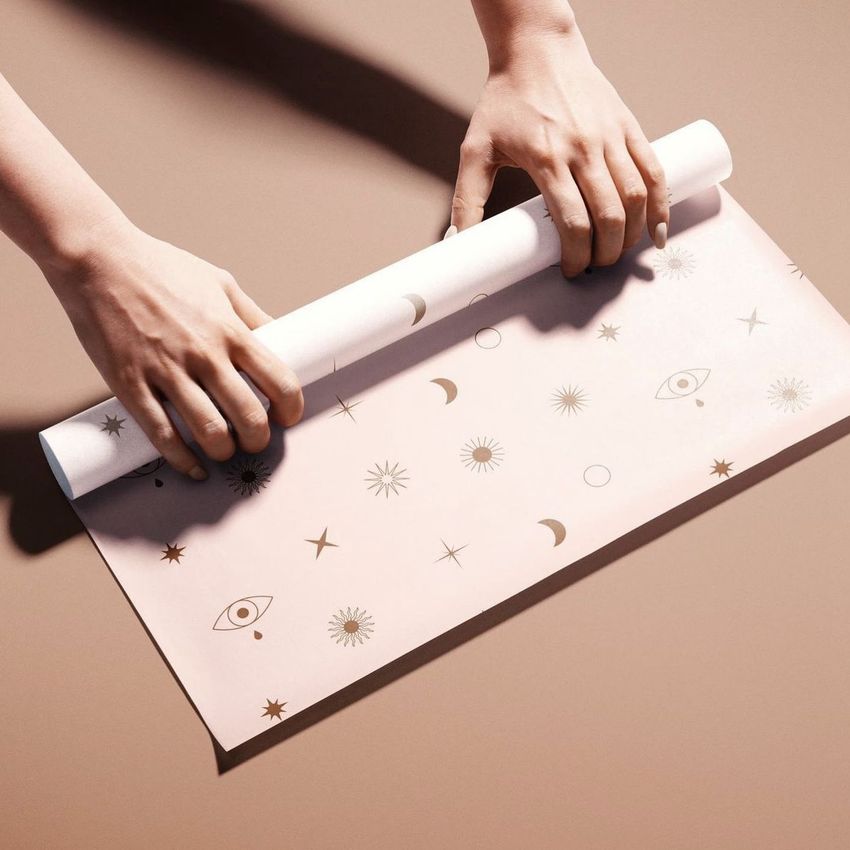 Hands unrolling a sheet of food safe paper mockup with gold celestial and eye illustrations on a beige surface.