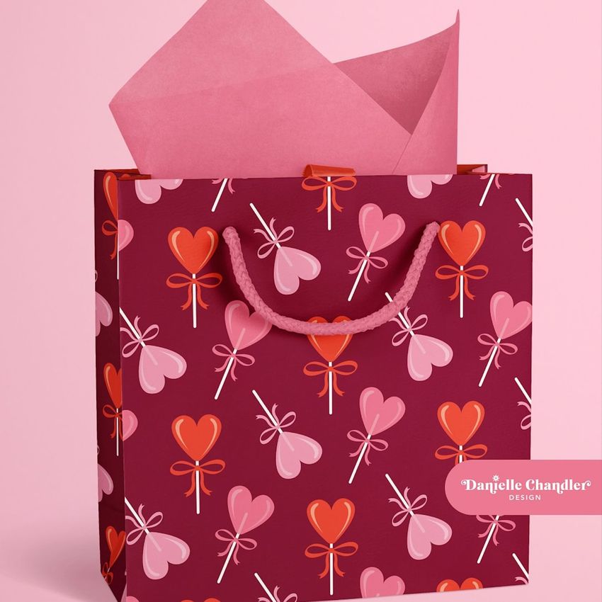 Gift bag mockup with pink heart-shaped lollipop pattern, pink rope handles, and pink tissue paper on a light pink background.