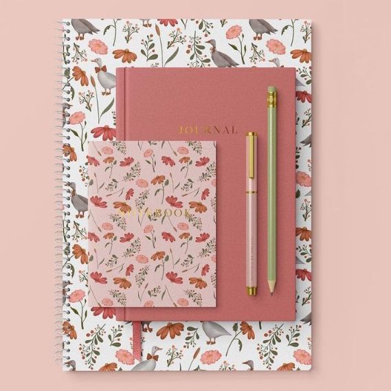 Stationery set mockup with a spiral notebook, journal, and notepad featuring geese and wildflowers pattern, arranged on a pink background.