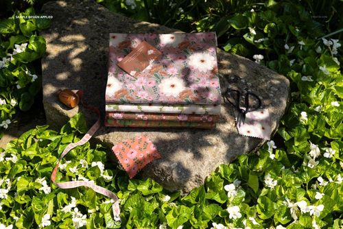 PSD mockup showing three folded fabric pieces with pink and white flower patterns on a stone in a lush garden setting.