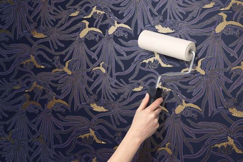 Paper wallpaper mockup showing a hand rolling out blue wallpaper with intricate gold floral motifs, front perspective.