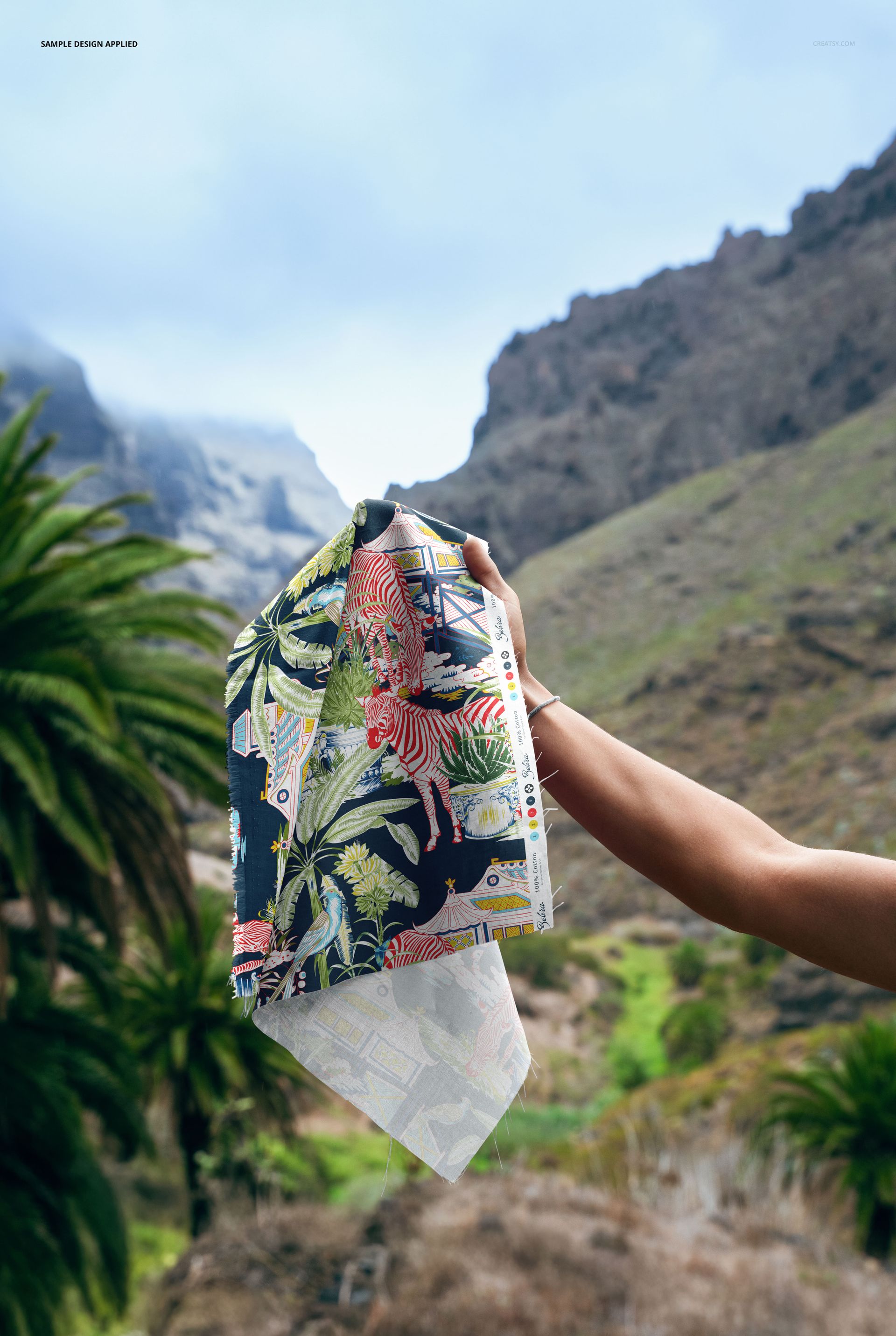 Tropical-themed fabric sample mockup held up outdoors, with vivid foliage design and scenic valley landscape behind.