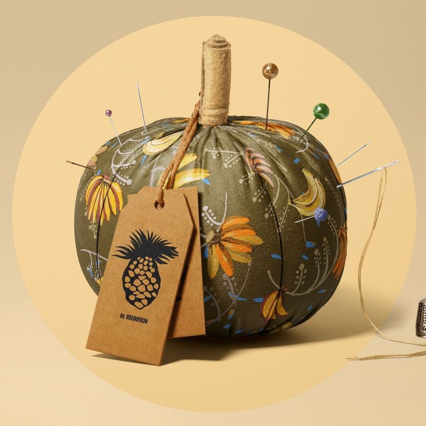 Fabric pumpkin mockup with a green floral pattern, used as a pincushion with sewing pins and a kraft tag on a beige background.