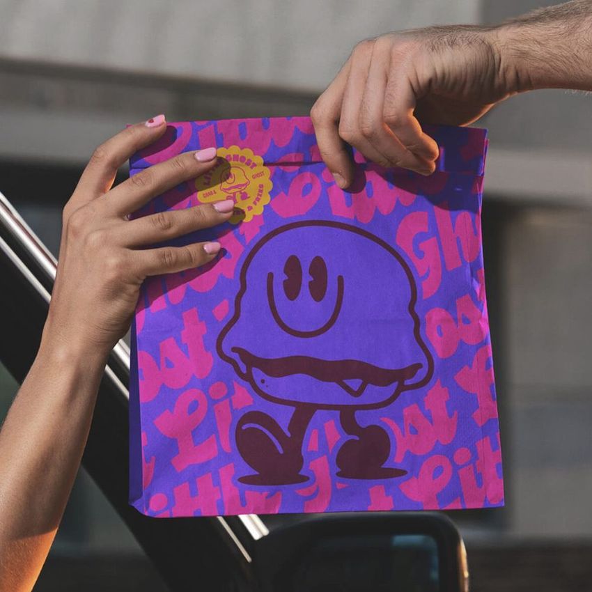 Fast food burger paper bag mockup featuring a bold purple cartoon ghost design and pink text pattern, held by two hands.