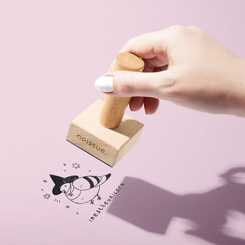 Wooden stamp mockup showing a hand stamping a playful drawing and stars onto a pink background, with branding on the stamp.