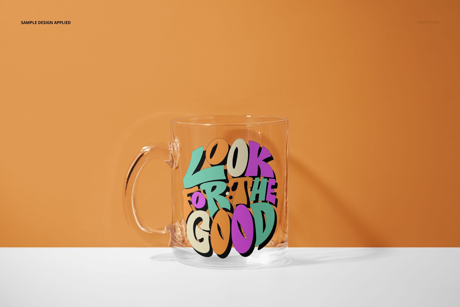 PSD mockup of a glass mug with multicolored text artwork, positioned on a white tabletop with an orange backdrop.