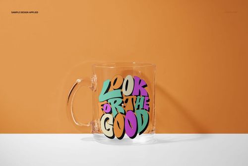 PSD mockup of a glass mug with multicolored text artwork, positioned on a white tabletop with an orange backdrop.