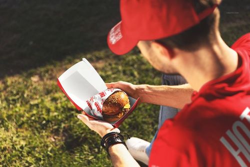 Person holding an open burger box with a wrapped burger inside, sitting outdoors on grass in sunlight.