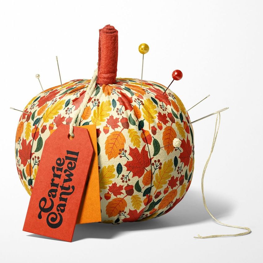 Fabric pumpkin mockup with a colorful autumn leaf pattern, red and yellow pins, and two tags attached to the stem on a white background.