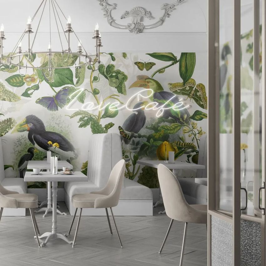 Cafe interior wallpaper mockup showing a botanical mural with tropical birds and leaves behind white tables and chairs.