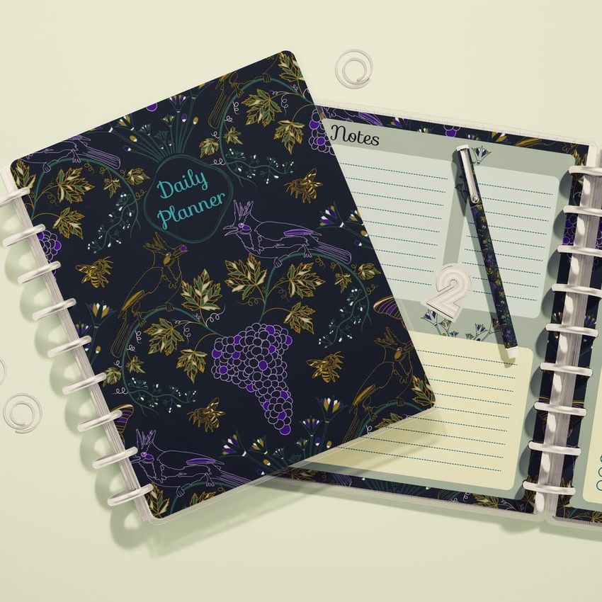 Top view of two spiral planners, one closed with a dark cover featuring gold and purple grape and bird illustrations, the other open to lined pages with a pen resting on top.