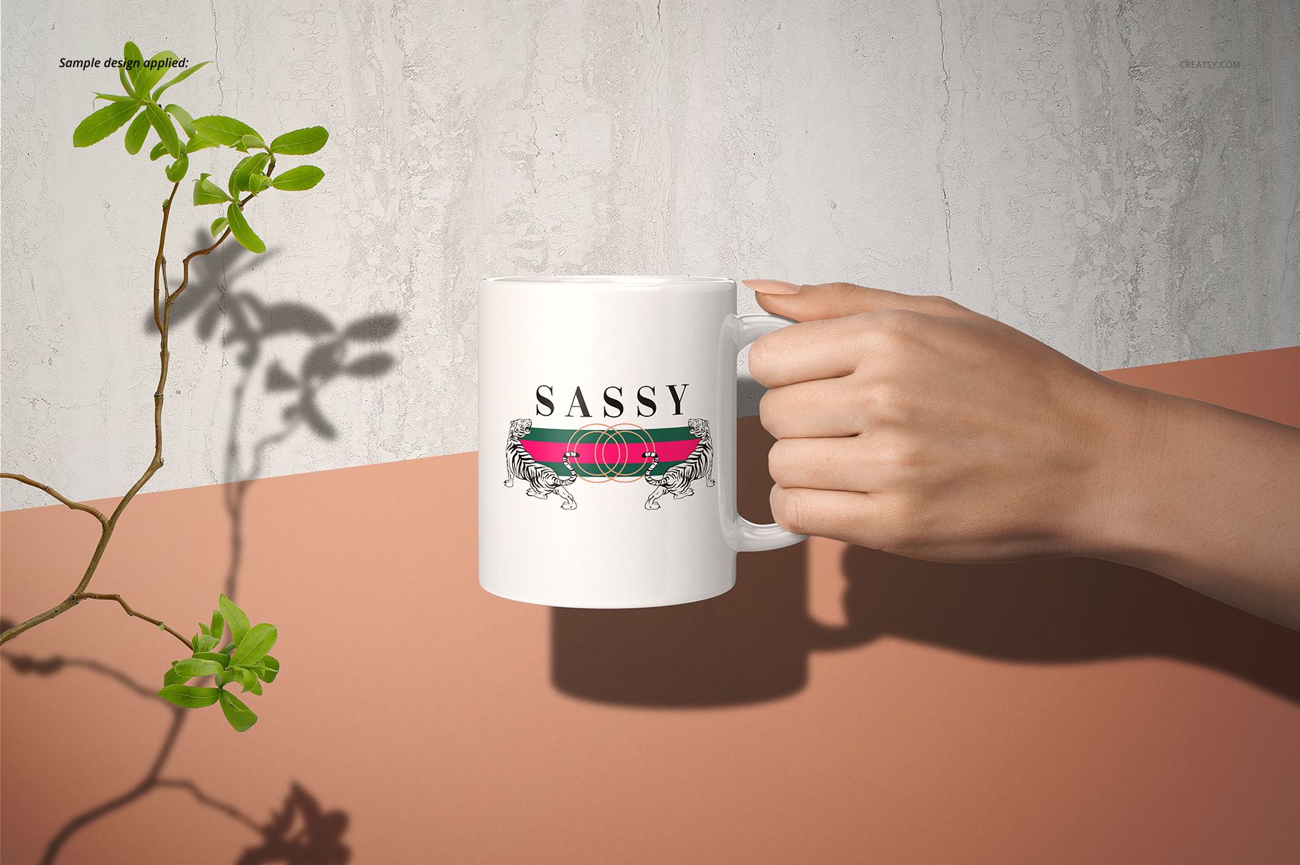 Hand holding a white 11oz mug with a bold text and decorative graphic, placed on a peach surface near a green plant.