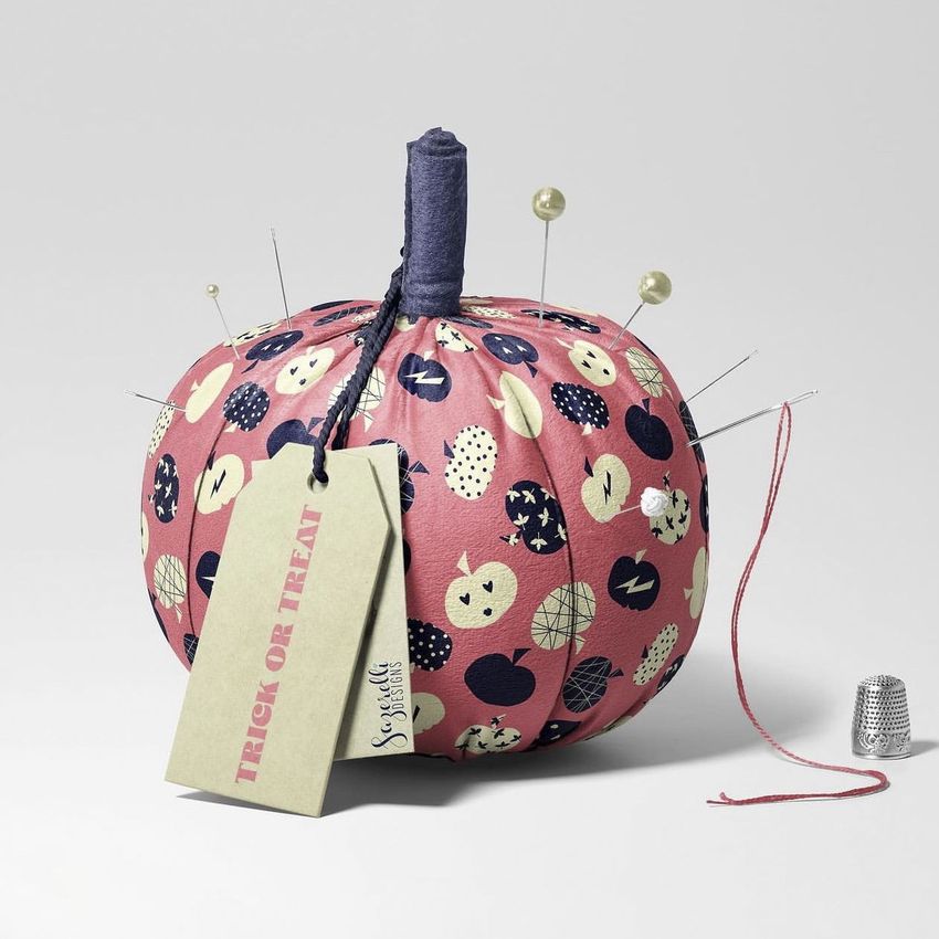Fabric pumpkin mockup featuring a pink pumpkin with a Halloween-themed pattern, sewing pins, a paper tag, and a metal thimble on a light gray background.