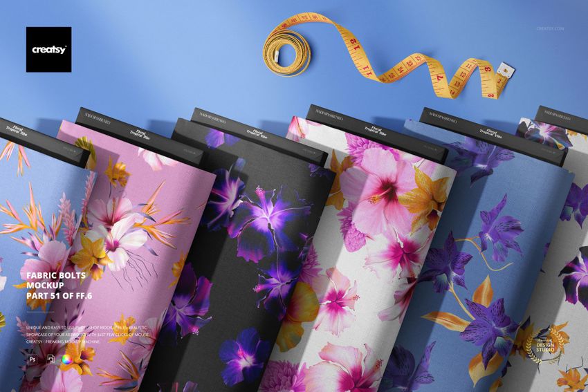 PSD mockup showing multiple fabric bolts featuring vibrant floral designs, displayed on a blue background with sewing tools.