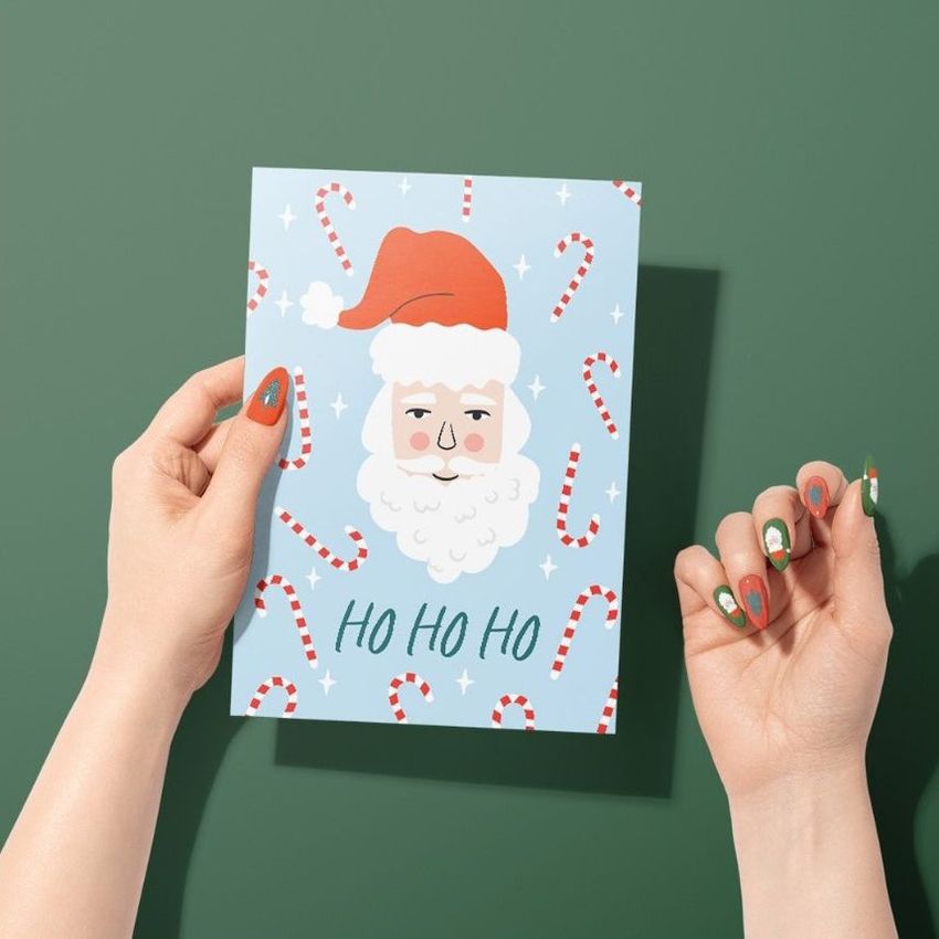 Greeting card mockup featuring a person holding a blue card with a Santa design and candy cane pattern against a green background.