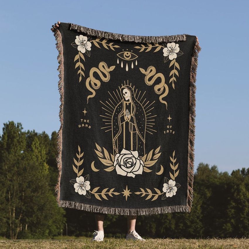 Woven throw blanket mockup held outdoors, featuring a gold and cream mystical illustration with floral and snake motifs.
