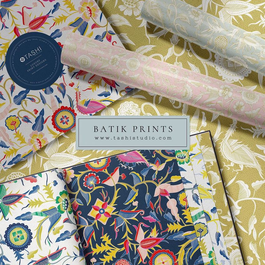 Wallpaper sample book mockup featuring open pages with colorful batik-inspired floral and bird patterns, alongside rolled wallpaper samples on a patterned surface.