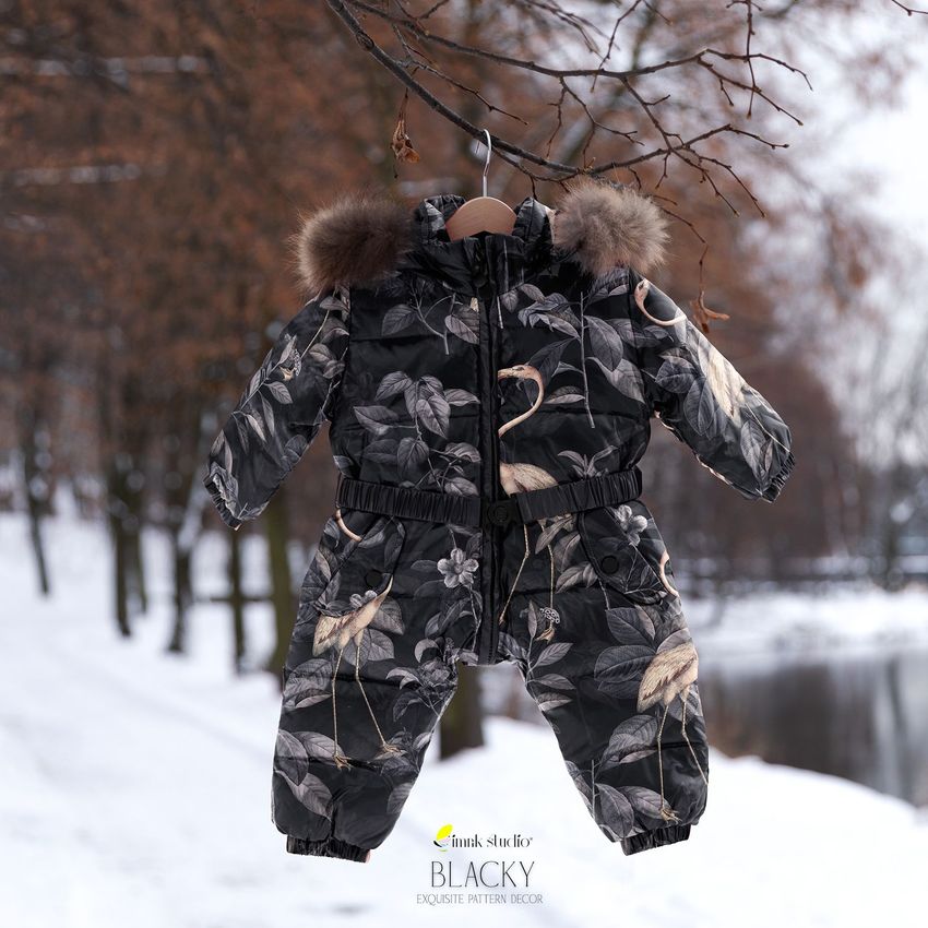 Baby snow suit mockup with a black floral and crane pattern, hanging on a wooden hanger outdoors in a snowy park.