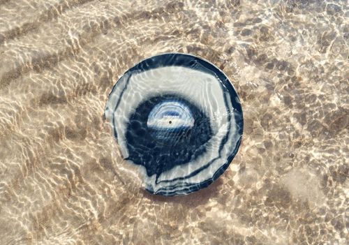 PSD mockup showing a round vinyl record with concentric color rings placed in shallow beach water, sunlight reflecting off sand.