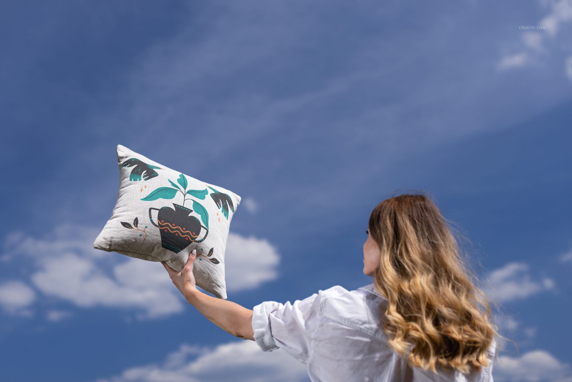 Woven pillow mockup with earthy pattern held up by a woman, with blue sky and clouds in the background.