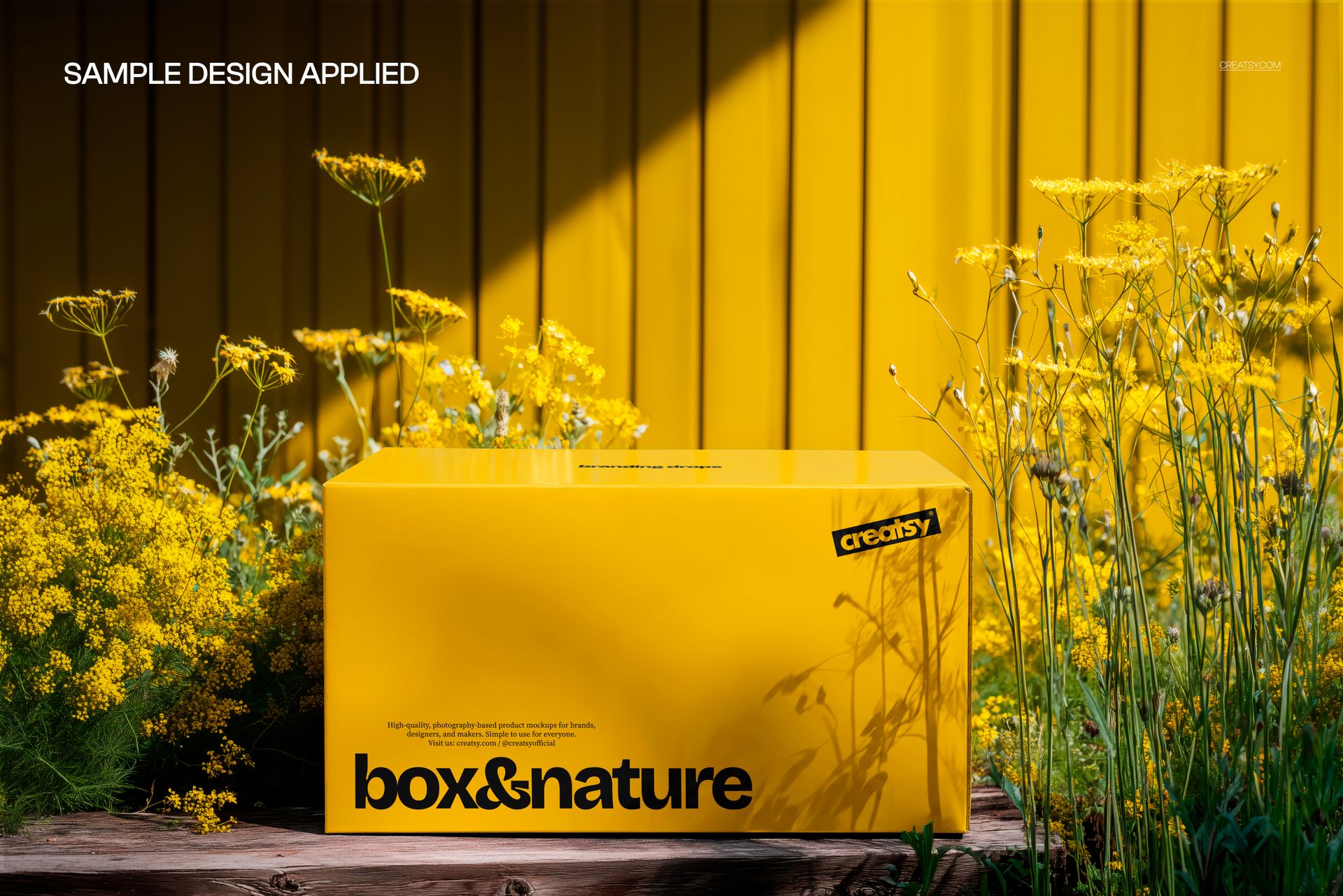Yellow rectangular box mockup placed among blooming wildflowers, viewed from the front against a yellow wall.