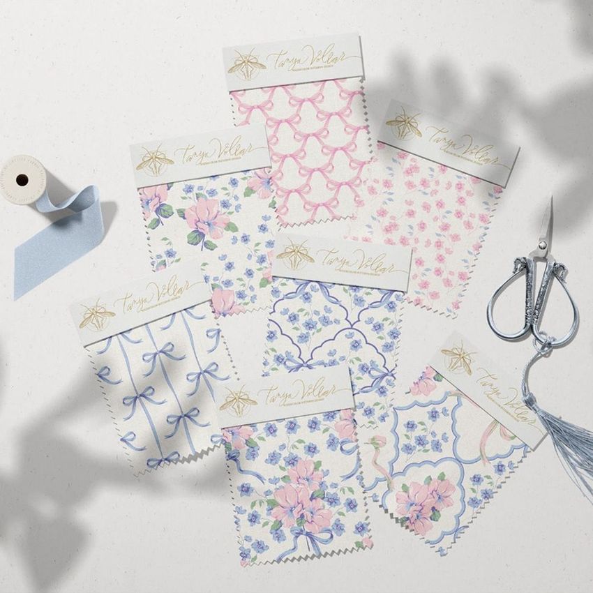 Fabric swatches mockup featuring six patterned fabric samples with floral and bow designs in pastel blue and pink, arranged on a light surface with scissors, ribbon, and a spool as props.
