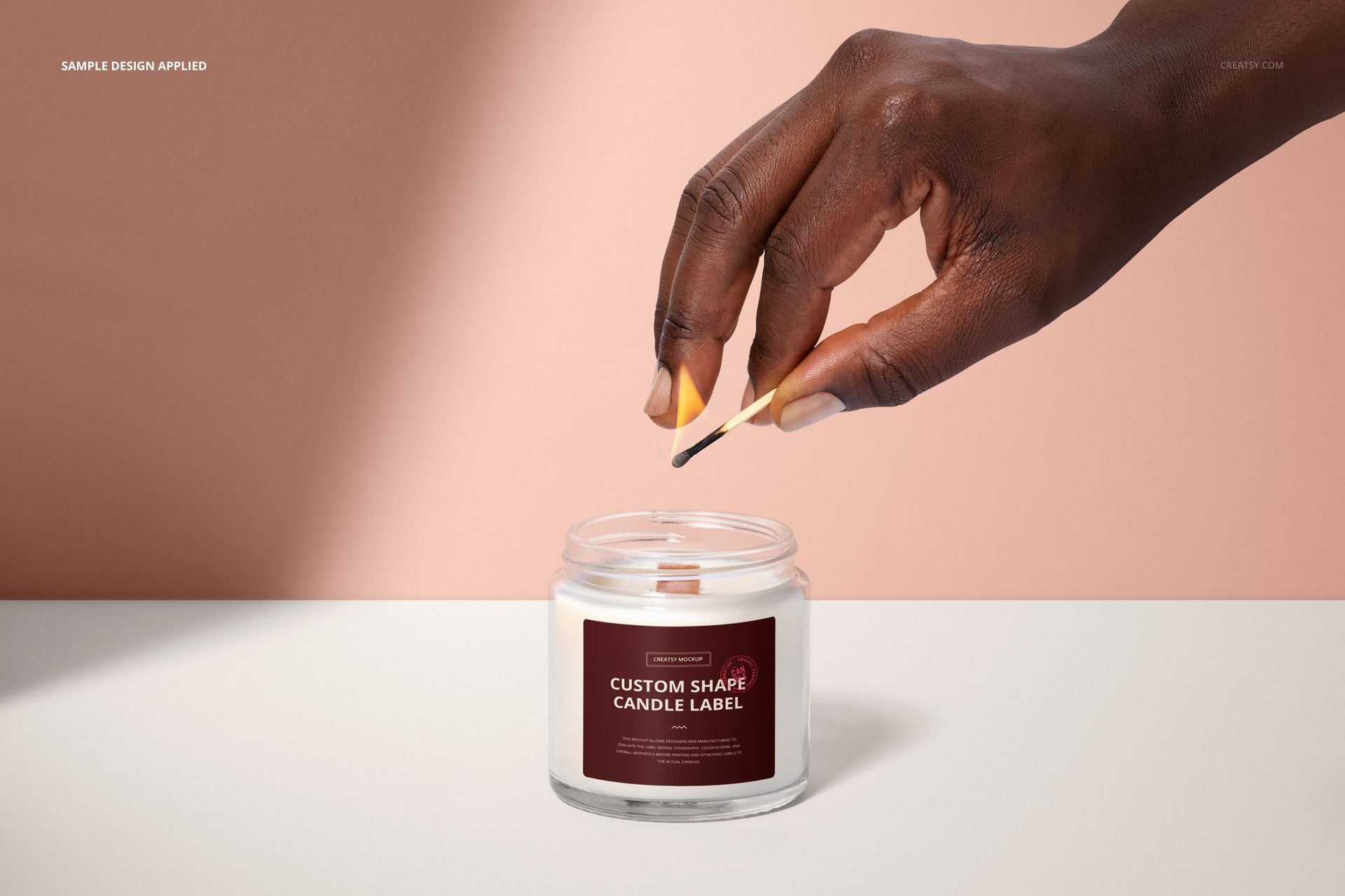 PSD mockup of a round glass candle jar with a colored label, being lit by hand, set on a white table and peach background.