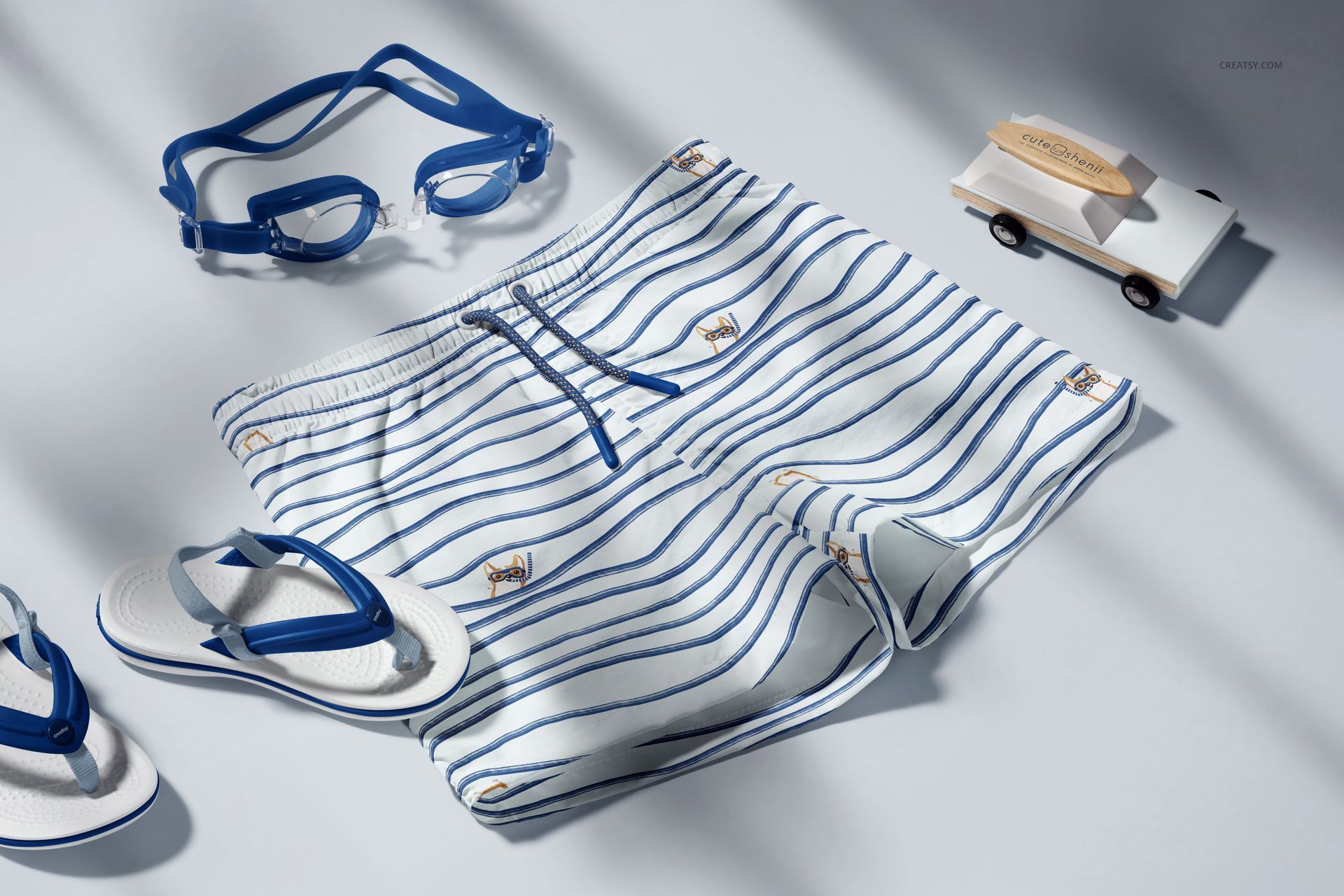 Striped kids swim shorts mockup shown from top view, accompanied by blue goggles, sandals, and a toy car.