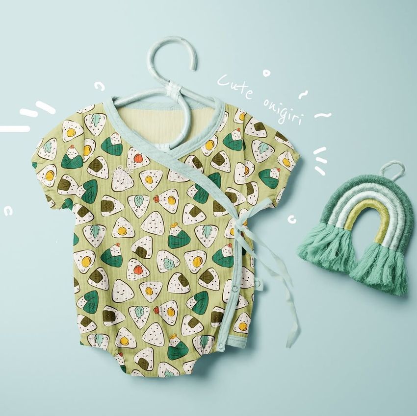 Muslin onesie mockup displayed on a hanger, featuring a playful onigiri pattern against a light blue background.