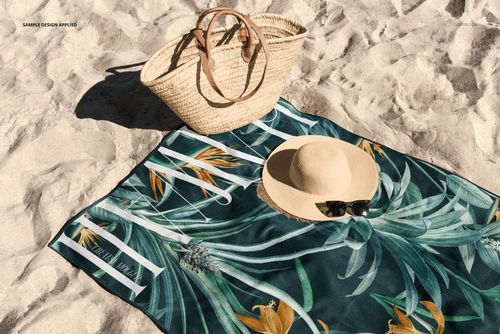 PSD mockup showing a rectangular beach towel with botanical print, straw bag, and sun hat on sand background.