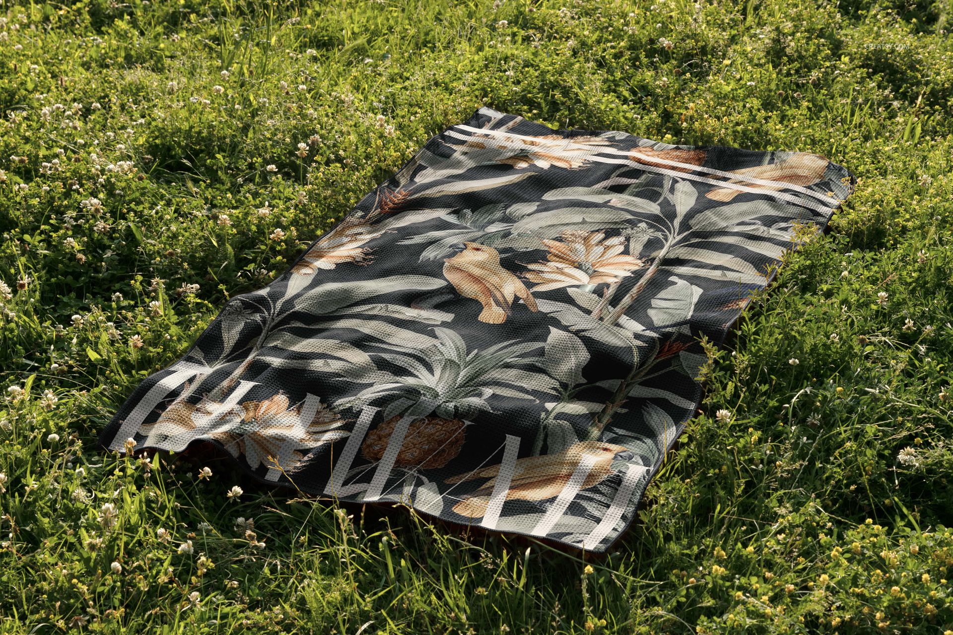 Outdoor scene showing a patterned waffle beach towel mockup spread on lush grassy ground with sunlight and shadows.