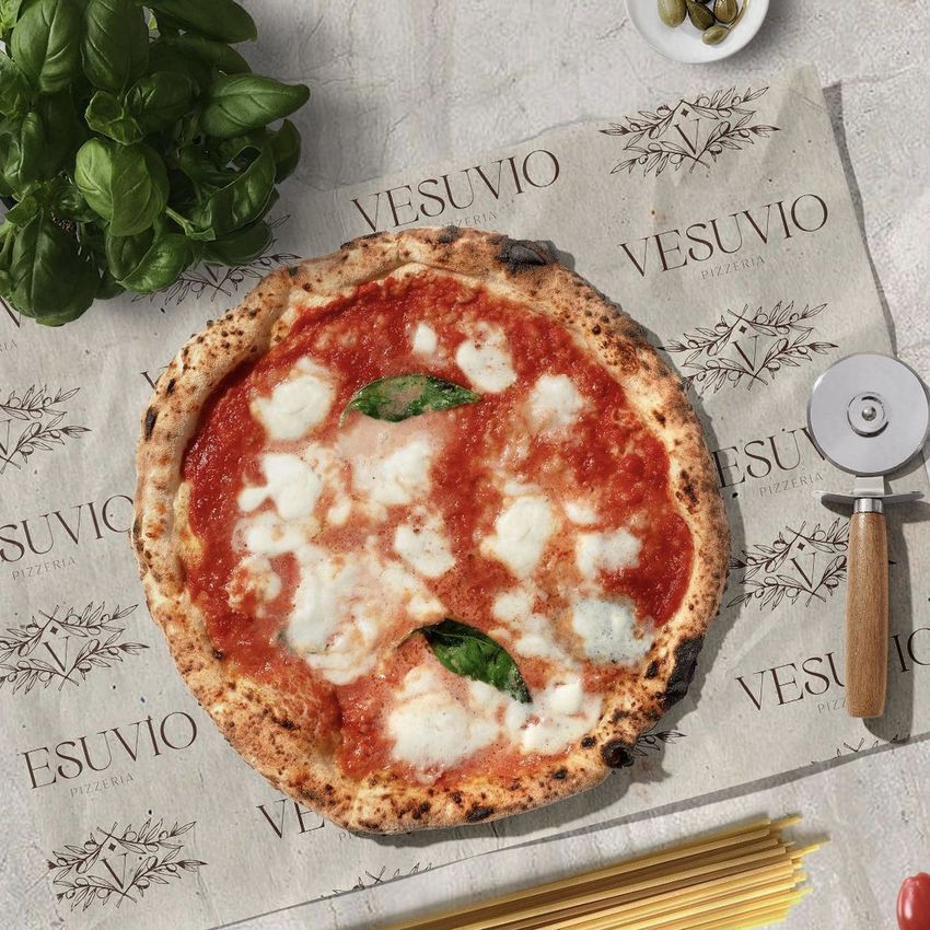 Pizza mockup on branded food safe paper with Vesuvio logo, surrounded by fresh basil, spaghetti, a pizza cutter, and olives on a light surface.