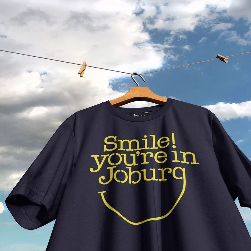 Navy blue t-shirt mockup hanging on a wooden hanger against a bright sky with clouds, featuring bold yellow text on the front.