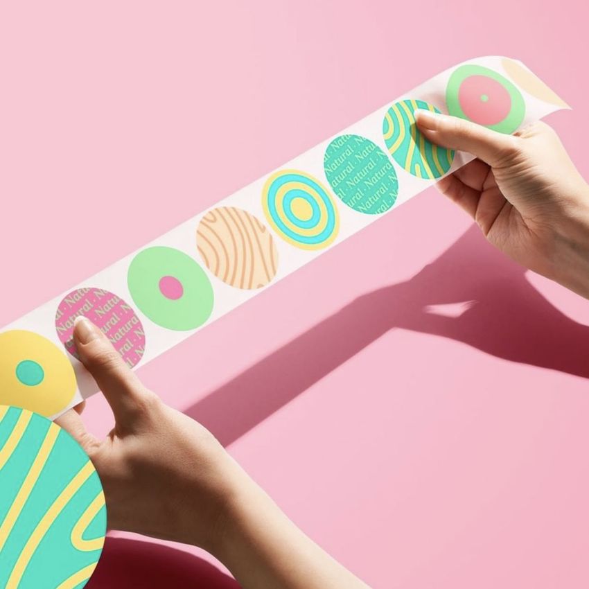 Close-up of hands unrolling a sticker mockup sheet with vibrant geometric and text-based designs on a pink backdrop.