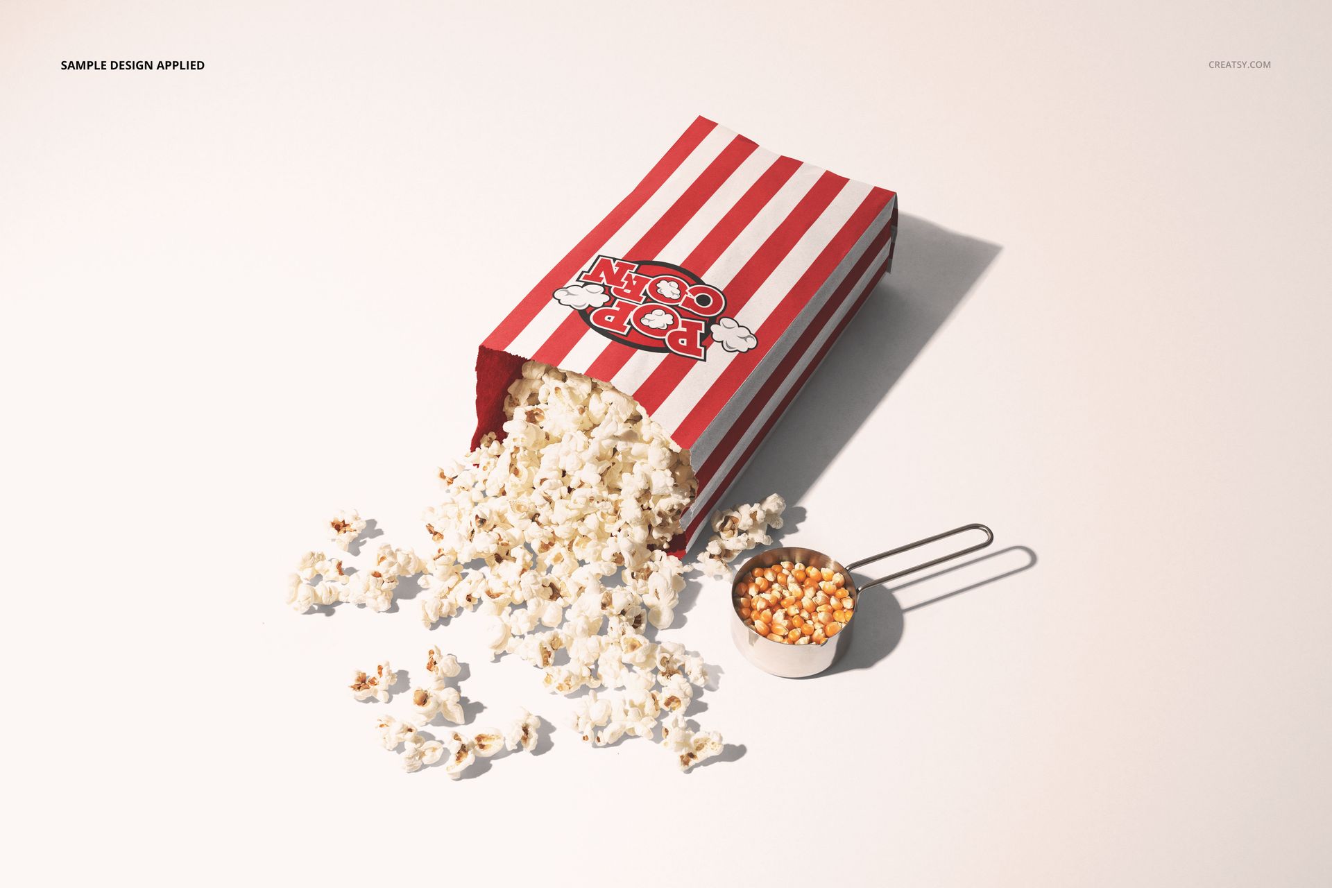 Red and white striped popcorn paper bag mockup shown from a side angle with popcorn spilling onto a light surface.