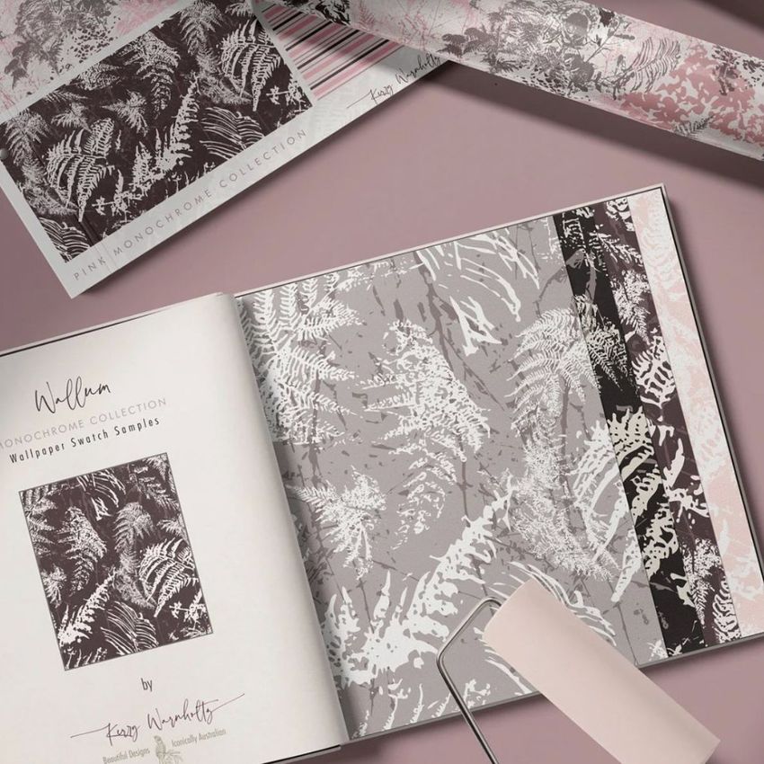 Wallpaper sample book mockup open to pages showing fern patterns in monochrome shades, with a paint roller and wallpaper roll nearby.