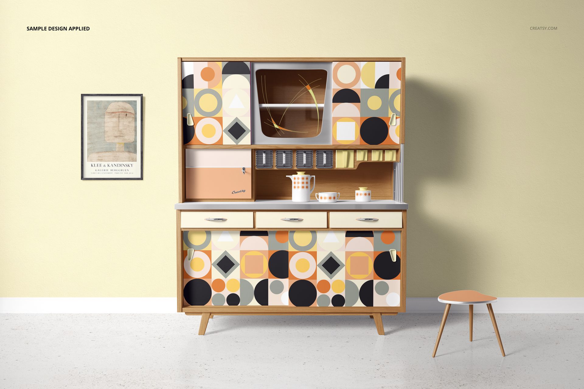 Front view of a kitchen cupboard mockup featuring colorful geometric designs, next to a small round stool and wall art.