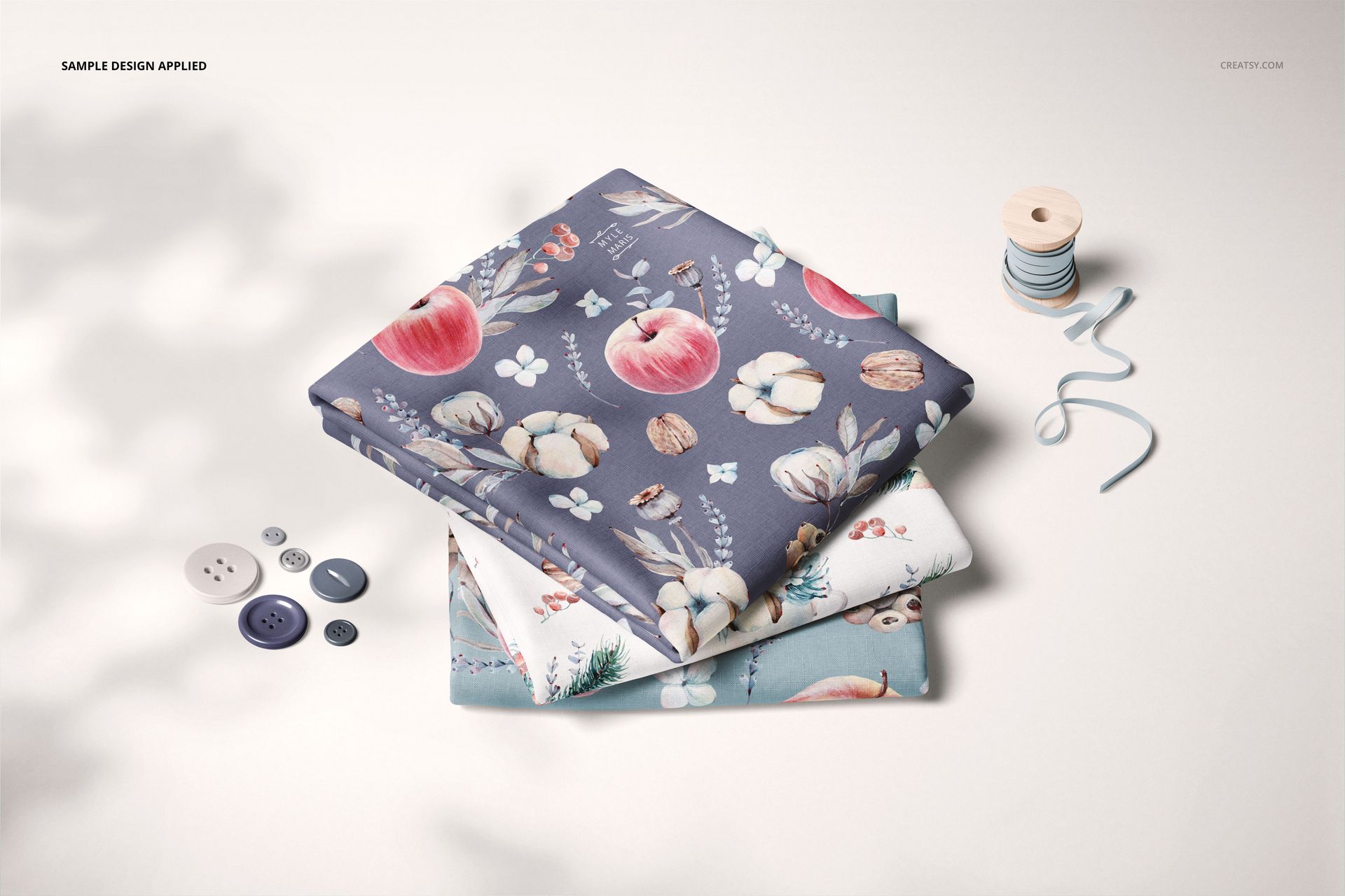 Folded fabric PSD mockup set showing three patterned textiles, surrounded by sewing accessories and soft shadows.