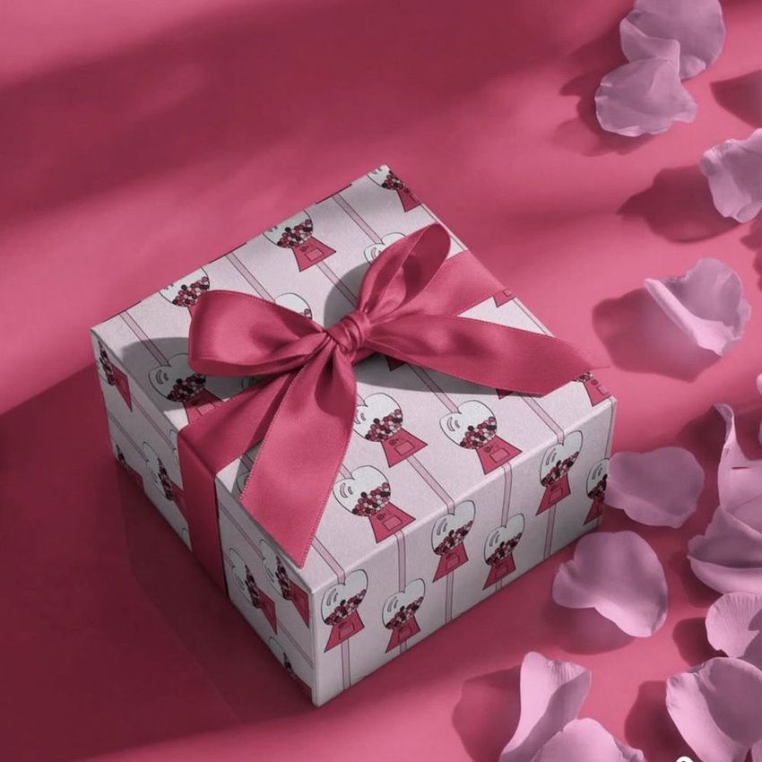 Square gift box mockup with a pink ribbon, decorated with cartoon illustrations, placed on a pink surface with scattered petals.