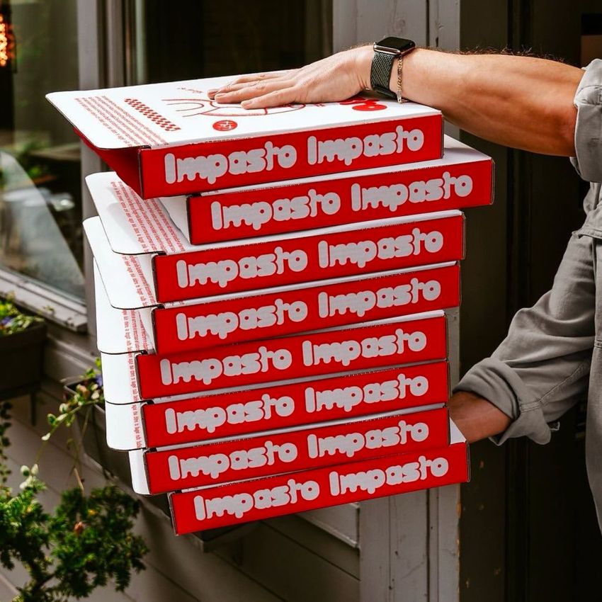 Stack of six pizza boxes with red and white design, held outdoors by a person in a gray shirt near a building window.